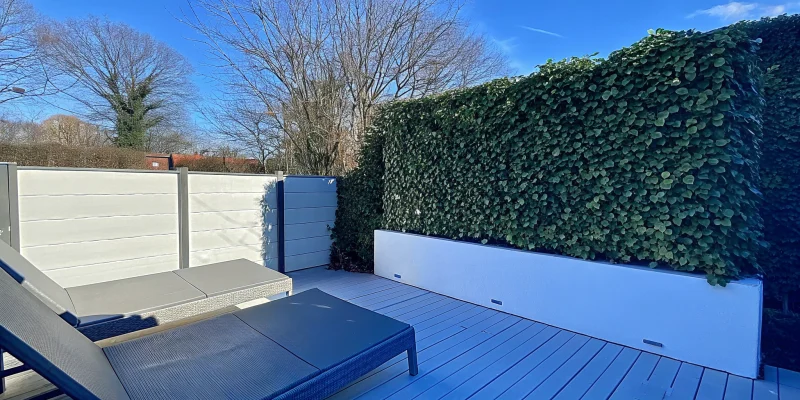 An outdoor artificial ivy wall on a white fence creates privacy for a modern patio.