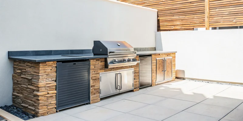 A modern stone veneered outdoor kitchen with a built-in grill and stainless steel appliances.